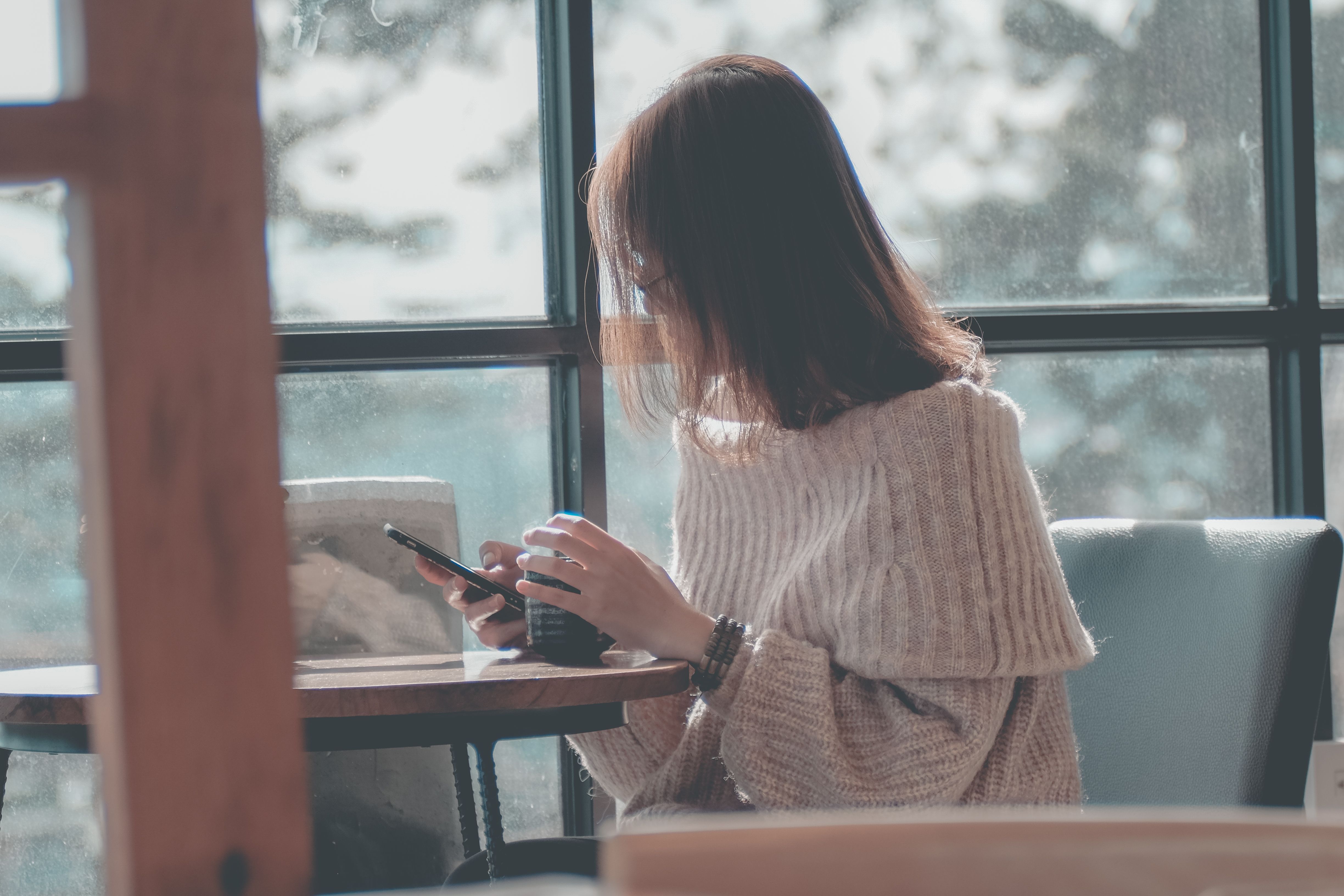 Una mujer mira su teléfono, sentada a una mesa junto a una ventana